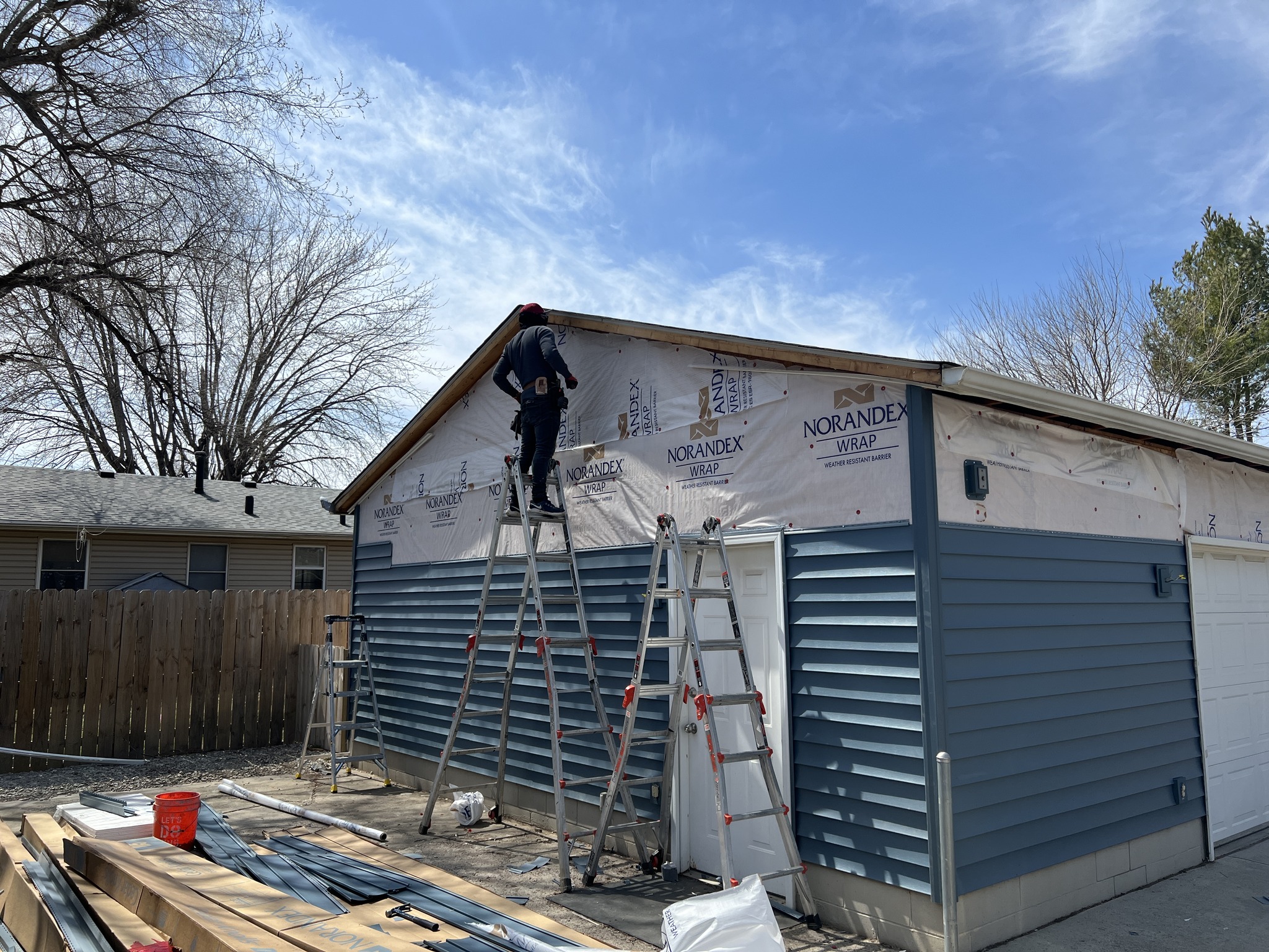 Residential blue siding installation in progress with ladders and house wrap.